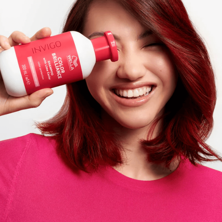Woman with vibrant red hair holding Wella Professionals Invigo Color Brilliance shampoo, smiling.